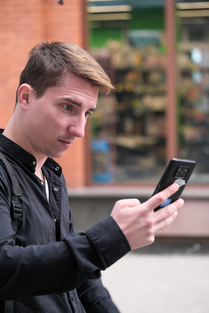 Portrait of a young handsome guy in a black shirt next to a brick wall with a smartphone autumn...