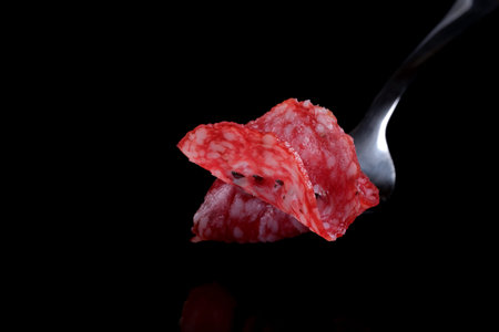 Thinly sliced delicious fragrant salami sausage on a steel fork close-up on a black mirror...