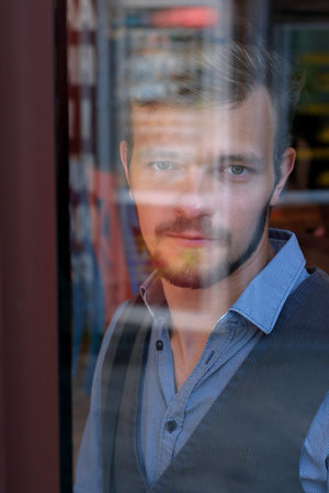 Street portrait of a young handsome man with a small beard and mustache in a cafe behind glass