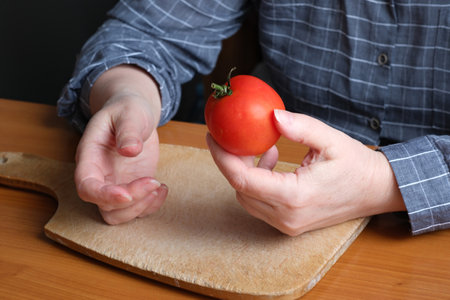 Slicing a red ripe tomato. An elderly woman's hands hold a tomato for slicing on a table in the kitchen. Close-up plan.の写真素材