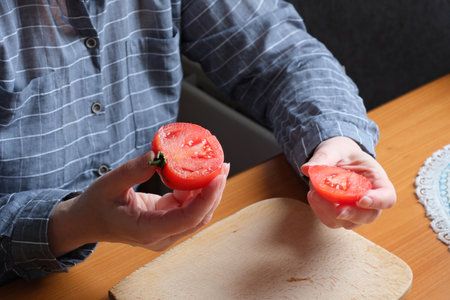 Slicing a red ripe tomato. An elderly woman's hands hold a tomato for slicing on a table in the kitchen. Close-up plan.の写真素材