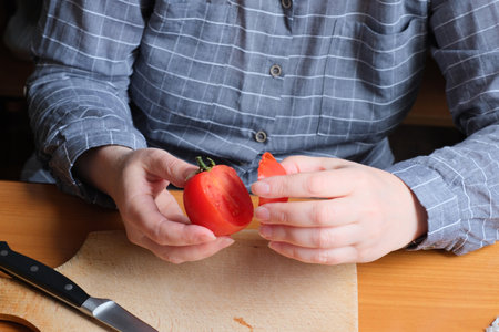 Slicing a red ripe tomato. An elderly woman's hands hold a tomato for slicing on a table in the kitchen. Close-up plan.の写真素材