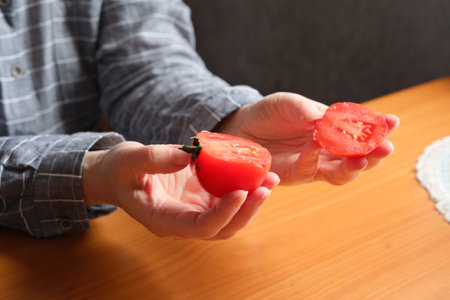 Slicing a red ripe tomato. An elderly woman's hands hold a tomato for slicing on a table in the kitchen. Close-up plan.の写真素材