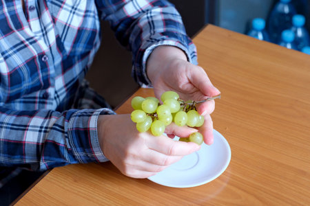The hands of an elderly woman holds a small brush of white seedless grapes and displays them in the kitchen at a brown table, without a face, close-upの写真素材