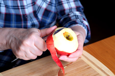 The hands of an elderly woman peel a red ripe tasty juicy apple with a knife in the kitchen at a brown table, without a face, close-upの写真素材