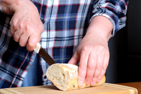 The hands of an elderly woman cut white wheat bread baguette with a knife in the kitchen at a brown...