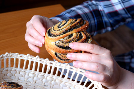 The hands of an elderly woman operate a sweet bun with poppy seeds in the kitchen at a brown table, without a face, close-upの写真素材