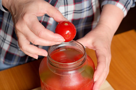 The hands of an elderly woman take out delicious homemade pickled tomatoes from a jar in the kitchen at a brown table, without a face, close-upの写真素材