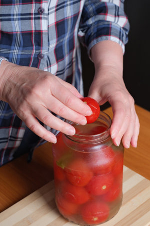 The hands of an elderly woman take out delicious homemade pickled tomatoes from a jar in the kitchen at a brown table, without a face, close-upの写真素材