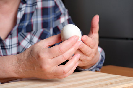 Hands of an elderly woman showing a white boiled egg and peeling it in the kitchen at a brown table, no face, close-upの写真素材