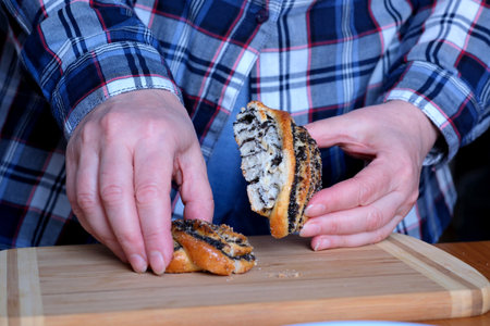 The hands of an elderly woman operate a sweet bun with poppy seeds in the kitchen at a brown table, without a face, close-upの写真素材