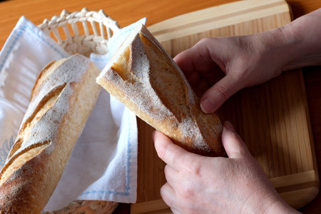 Hands of an elderly woman holding sliced white wheat bread baguette in the kitchen at a brown table, no face, close-upの写真素材