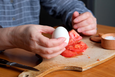 Hands of an elderly woman showing a white boiled egg and peeling it in the kitchen at a brown table, no face, close-upの写真素材