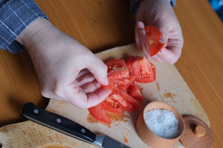 Hands of an elderly woman cutting a red ripe tomato on a cutting board for a snack in the kitchen at a brown table, no face, close-upの写真素材