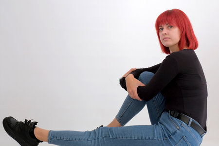 Beautiful cute young woman with orange hairstyle in jeans sitting posing in studio on white backgroundの写真素材