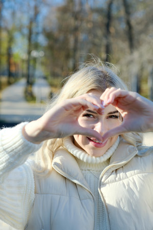 Portrait of a young beautiful blonde with long hair in a white sweater and jacket in sunny autumn in a city parkの写真素材