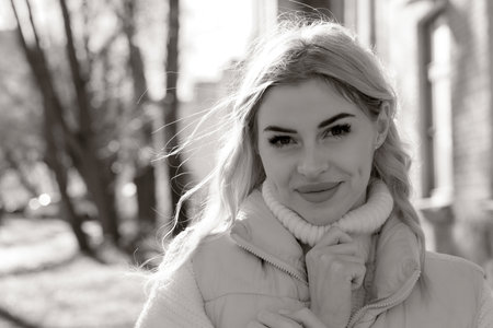Portrait of a young beautiful blonde with long hair in a white sweater and jacket in sunny autumn in a city park black and white imageの写真素材