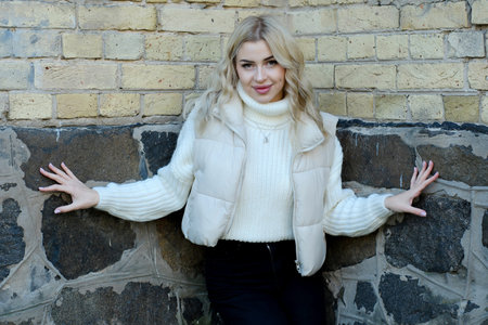 Portrait of a young beautiful blonde with long hair in a white sweater and jacket in a brick cornerの写真素材