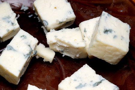 Mouthwatering blue cheese squares are neatly cut and displayed on a textured wooden platter, showing their creamy, crumbly texture and blue veining. A visual feast for cheese lovers.の写真素材