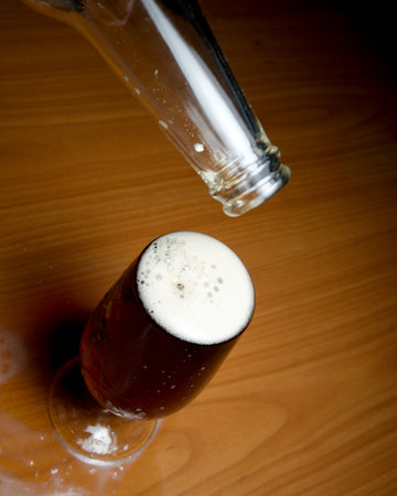 Foamy amber drink cascades into a stylish glass on a wooden table.の写真素材