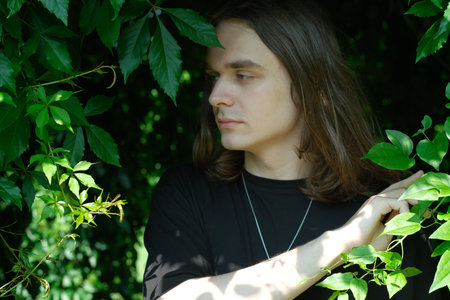 A young man with long hair is peering through vibrant green foliage, captivated by nature.の写真素材