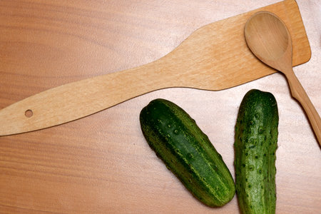 Two crisp cucumbers lie beside a wooden spatula and spoon on a warm kitchen surface.の写真素材