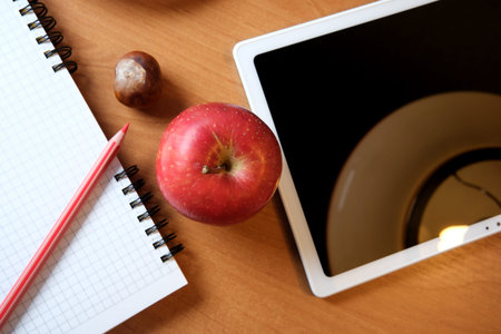 Red apple, notebook, and tablet arranged on a warm wooden table in sunlight.