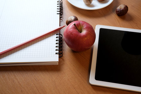 A red apple sits next to a tablet and an open notebook on a wooden table.