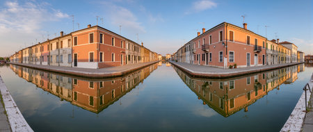 Colorful buildings line a serene canal, mirrored in calm waters under a clear sky.の素材