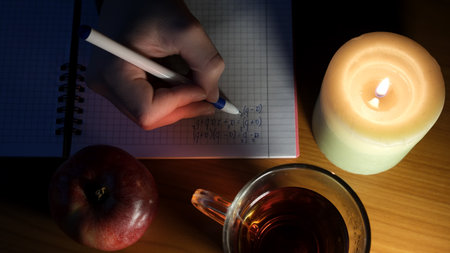 A student writes notes by candlelight, enjoying tea and an apple during study time.の写真素材