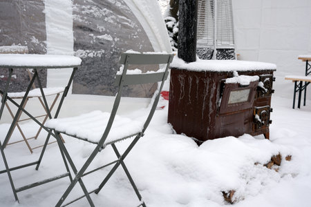 Chairs covered in snow sit near a stove, creating a cozy winter scene outdoors.の写真素材
