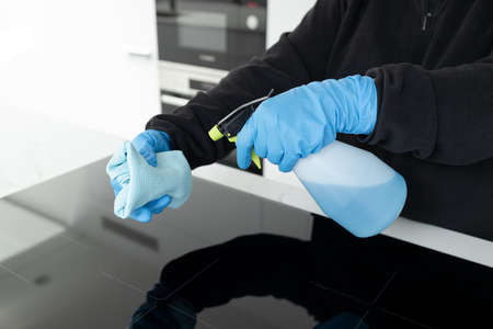 Hands in protective glove cleaning a modern electric stove with rag and spray of a minimalist kitchenの写真素材