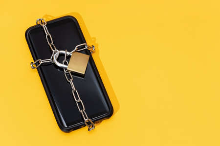 Phone security concept. Smartphone locked with chain and padlock on yellow background. Mobile security and data privacy. Copy spaceの写真素材