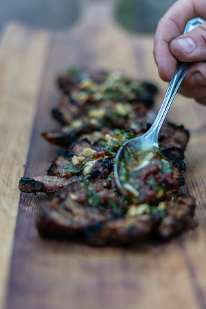 Hand placing sauce on grilled meat on a cutting wood with a spoon.の写真素材