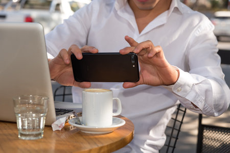 Man sitting outdoors using his smartphone while enjoying a cup of coffee in front of his laptopの写真素材