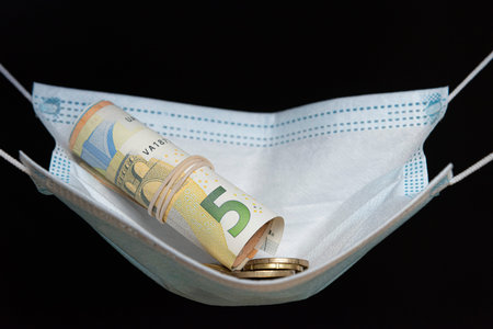 Medical face mask holding money inside of it, on a black background. World coronavirus economic...