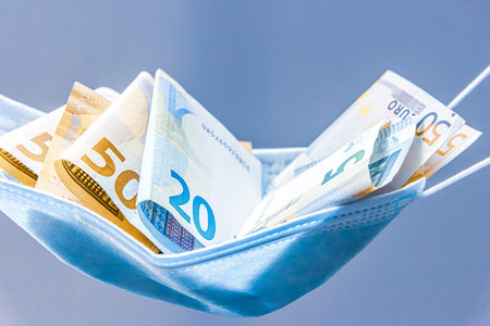 Medical face mask holding money inside of it, on a light background. World coronavirus economic...