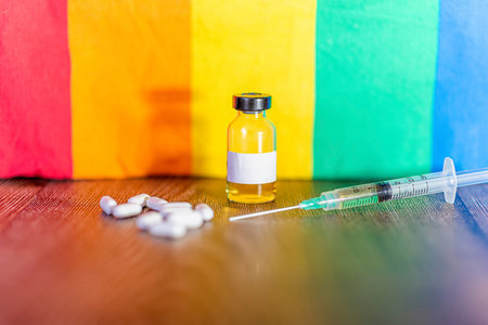 Syringe, vaccine vial and medicinal tablets in the background of a pride rainbow flag.