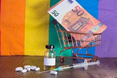 Injection, vaccine vial and medicinal tablets, Euro banknotes in a shopping cart. Background of a pride rainbow flag.の写真素材