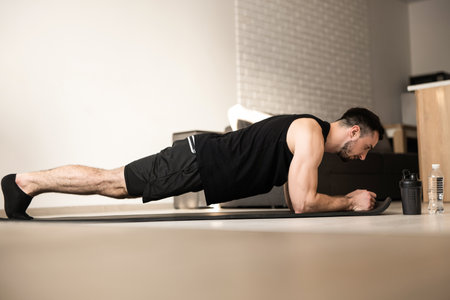 Plank it Training at home. Confident young man wearing black sportswear and doing plank position while exercising on the floor in light loft interior. Hard workout.の写真素材