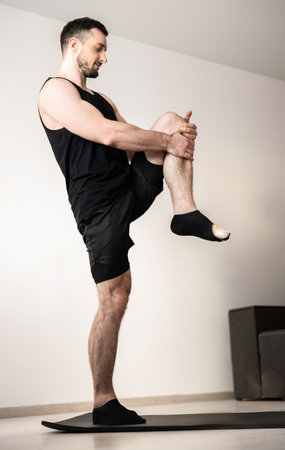 Athletic man standing on one leg and stretching another. Workout on black yoga mat. Muscular young man practicing yoga position at home. Morning warm up. Black sportswear. Light room on background.の写真素材