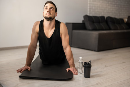 Fitn man meditating while doing cobra pose in living room. Indoor shot of handsome young man practicing yoga. Morning workout. Black sportswear. Plastic water bottle. Strong arms.の写真素材