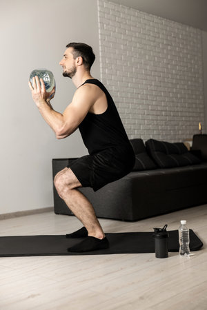 Fit man doing squat exercises with big bottle of water in hands at home. Apartment with minimalistic interior on background. Black sportswear. Healthy lifestyle, wellbeing and activity concept.の写真素材