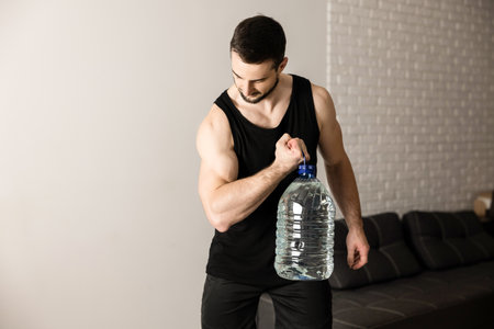 Athletic man doing calf raise exercises with big bottle of water. White modern room on background. Strong man in black sportswear excercising to have a fit body.の写真素材