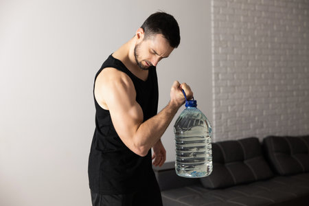 Strong man is doing calf raise exercises with big bottle of water at home in his spacious and bright apartment with minimalistic interior. Man in black sportswear doing morning workout.の写真素材