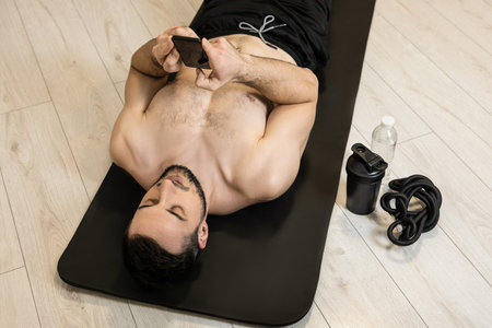 Fit man is resting after hard fitness and texting with friends on the phone. Sports equipment. Resting time. Lazy man lying on black yoga mat and messaging by smartphone.の写真素材