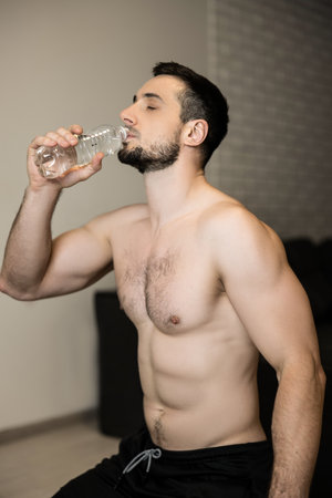 Topless attractive man drinking water after hard workout at home. Resting time. Sporty muscular man drinking water after exercising. Bodybuilding workout. Hydration and fitness concept.の写真素材