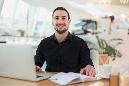 Cheerful freelancer man working from cafe. Attractive young man smiling, using his modern laprtop and looking at camera. Cozy coffeehouse on background. Blurred open diary, mug of coffee on table.の写真素材
