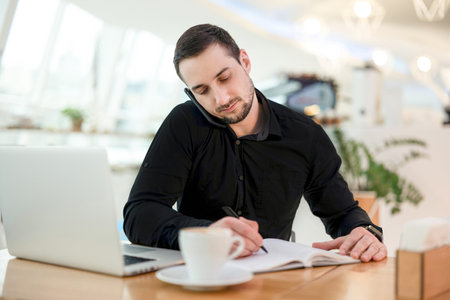Businessman writes date of meeting with client in his planner. Young man in black shirt working from coffeehouse, drinking cappuccino and using his modern laptop.の写真素材