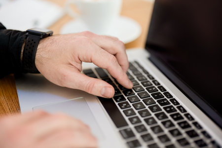 Male hands typing on laptop keyboard. Cut view. Modern gadgets. Close up. Gray laptop. Blurred cup of coffee on background. Man in black shirt. Male freelancer working from cafe.の写真素材